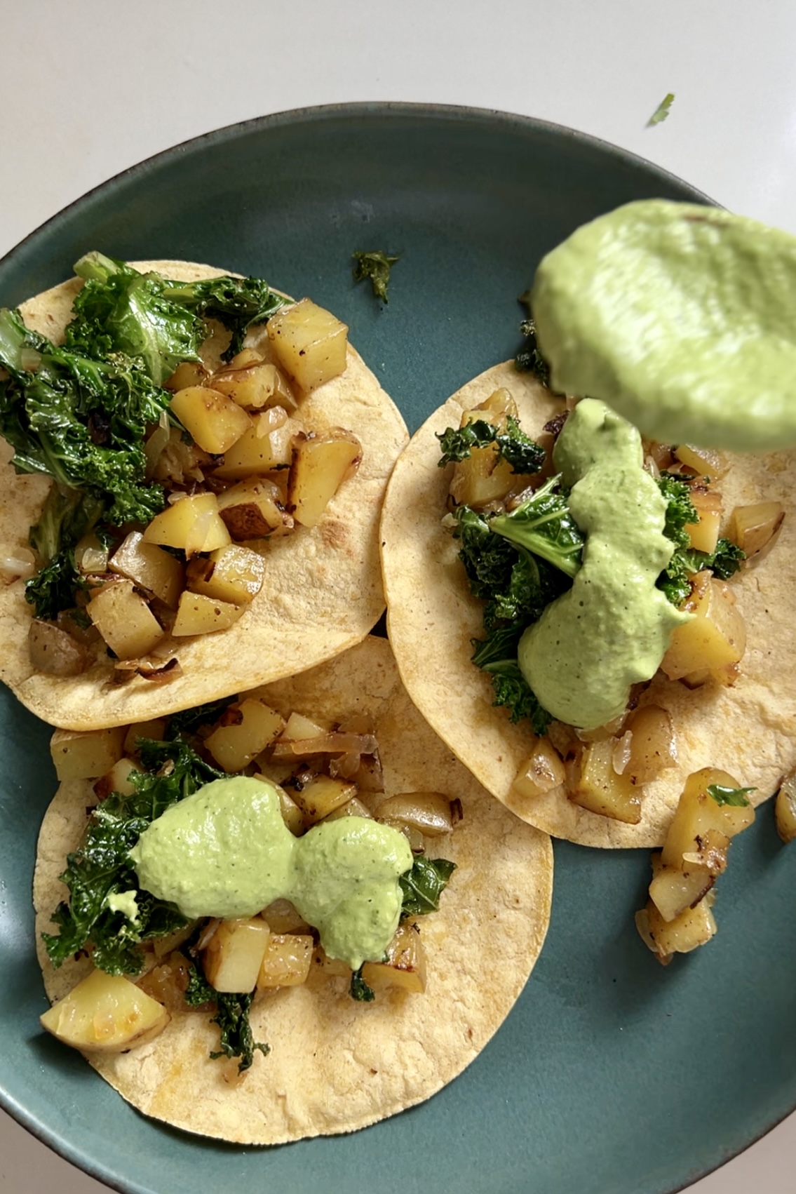 A plate with three potato tacos on a teal ceramic plate. Each taco consists of sautéed diced potatoes and wilted kale on small corn tortillas. A spoonful of creamy green Cilantro Bitchin Sauce is being drizzled over the tacos, with some sauce already visible on each. The white background enhances the food's colors.