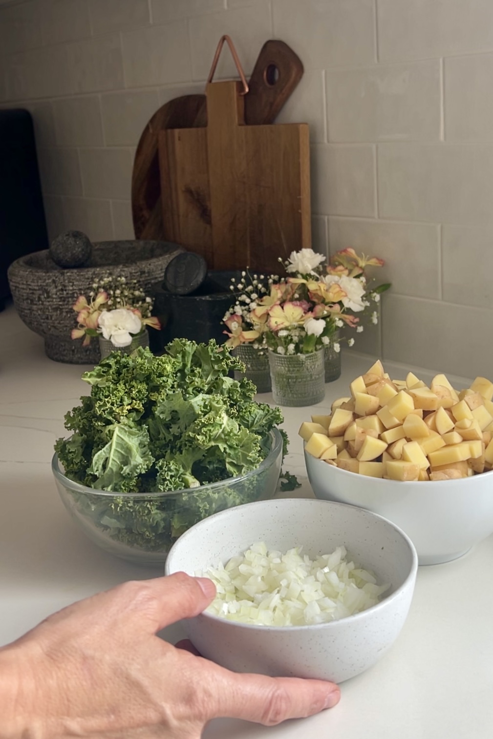 A hand holding a white bowl of chopped onions in the foreground, with other prepared ingredients including diced potatoes for kale potato tacos in a white bowl and kale in a glass bowl. The background features a tiled backsplash, elegant flowers, and wooden cutting boards arranged upright.