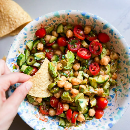 A hand is shown dipping a corn tortilla chip into a colorful, vegan ceviche salad in a bowl. The salad consists of chickpeas, cherry tomatoes, sliced cucumbers, olives, and greens, all mixed with a light dressing. The bowl has a blue and white speckled pattern. Another tortilla chip lies nearby.