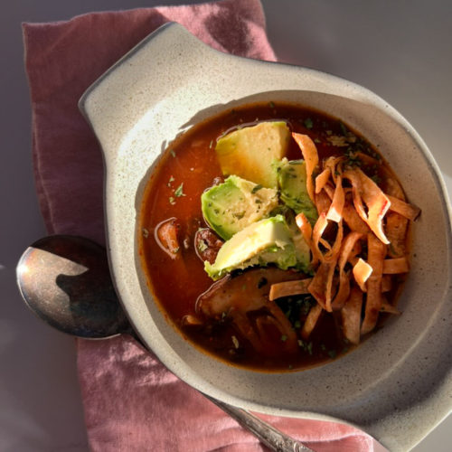 A white ceramic bowl filled with a vibrant red Mexican soup garnished with sliced avocado, chopped cilantro, and crispy tortilla strips rests on a pink cloth napkin. To the left of the bowl, a tarnished spoon casts a shadow on the napkin. The setting is illuminated by natural light.