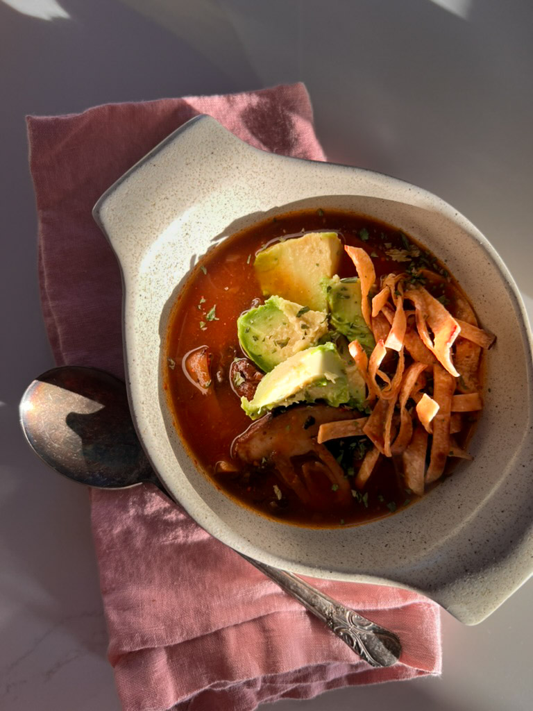A white ceramic bowl filled with a vibrant red Mexican soup garnished with sliced avocado, chopped cilantro, and crispy tortilla strips rests on a pink cloth napkin. To the left of the bowl, a tarnished spoon casts a shadow on the napkin. The setting is illuminated by natural light.