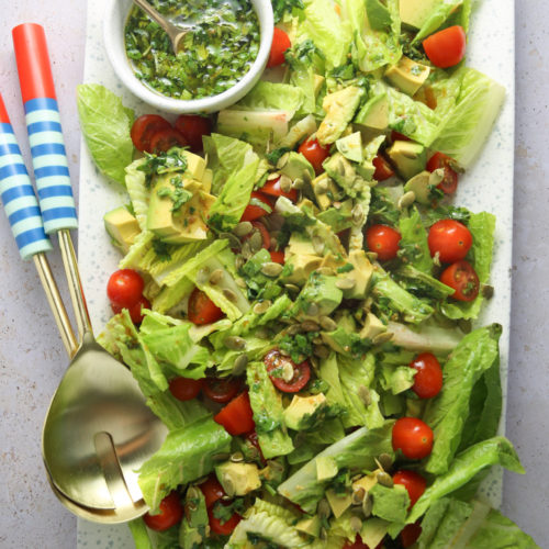 A vibrant salad is served on a rectangular white plate. The salad features chopped romaine lettuce, halved cherry tomatoes, and avocado pieces, garnished with seeds. A small bowl of cilantro dressing sits at the top left with two blue and white striped-handled spoons placed beside it.