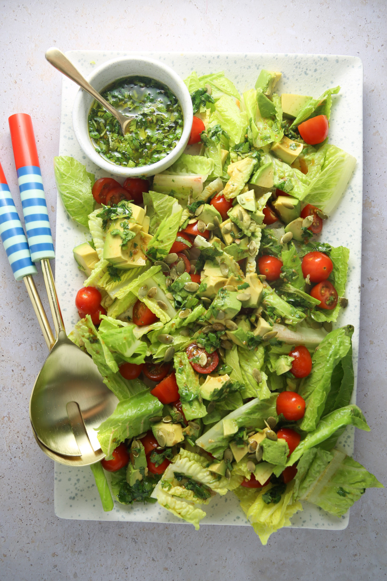 A vibrant salad is served on a rectangular white plate. The salad features chopped romaine lettuce, halved cherry tomatoes, and avocado pieces, garnished with seeds. A small bowl of cilantro dressing sits at the top left with two blue and white striped-handled spoons placed beside it.