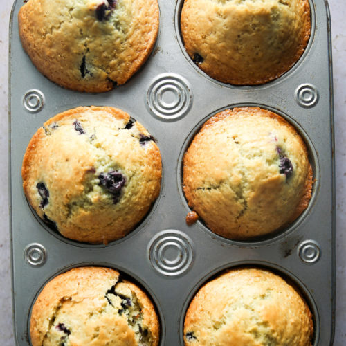 A close-up of six freshly baked Lemon Blueberry Muffins in a silver muffin tin resting on a light-colored countertop. The muffins are golden brown with visible blueberries and hints of lemon, appearing to be perfectly baked from scratch, with rounded, slightly cracked tops.