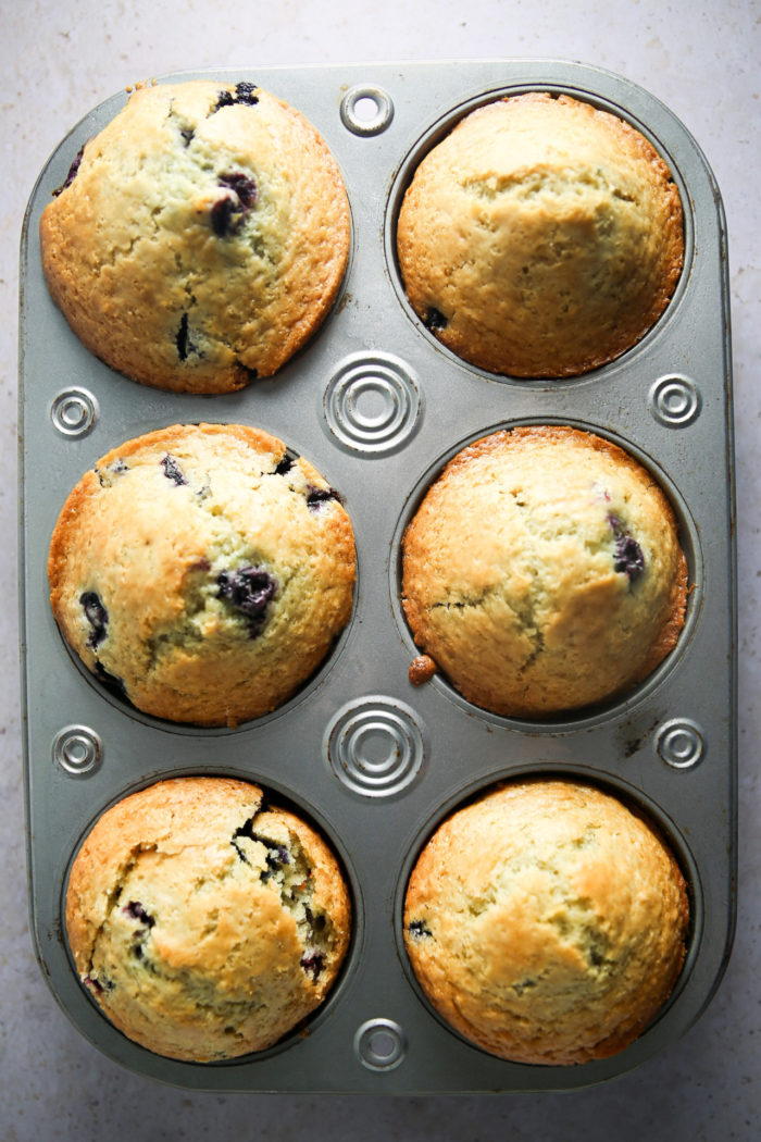 A close-up of six freshly baked Lemon Blueberry Muffins in a silver muffin tin resting on a light-colored countertop. The muffins are golden brown with visible blueberries and hints of lemon, appearing to be perfectly baked from scratch, with rounded, slightly cracked tops.