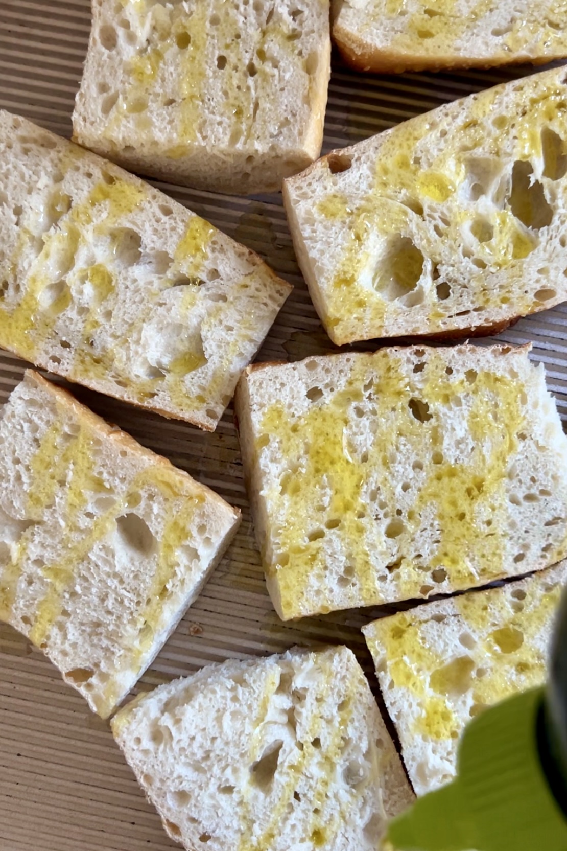 A close-up of several slices of ciabatta bread on a wooden surface, perfect for Pan Con Tomate. The bread is lightly drizzled with olive oil, giving it a yellowish tint and enhancing its porous and airy texture. The irregular holes and the soft, rustic appearance are characteristic of ciabatta.