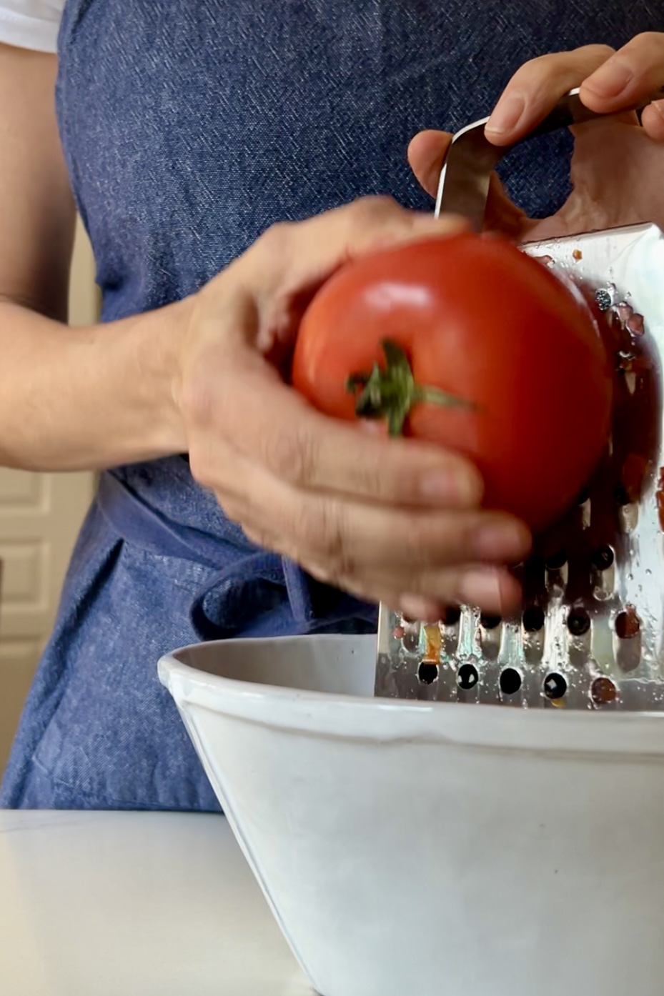 A close-up of a person wearing a blue apron grating a red tomato over a white container, perfect for preparing Pan Con Tomate. The person holds the tomato with their left hand while pressing it against the grater with their right hand. The image focuses on the hands, the tomato, and the grater, with the background slightly blurred.