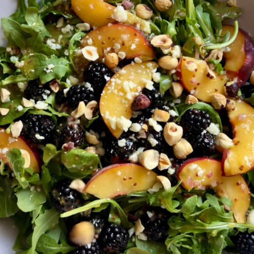 A fresh arugula salad in a white bowl featuring sliced peaches, blackberries, and crumbled cheese, topped with chopped hazelnuts. The colorful ingredients create a visually appealing combination, highlighting the vibrant mix of green, yellow, dark purple, and light brown hues.