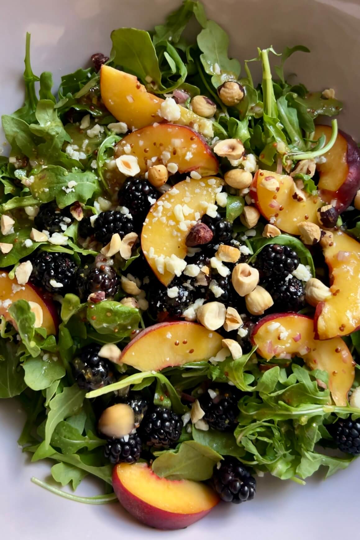 A fresh arugula salad in a white bowl featuring sliced peaches, blackberries, and crumbled cheese, topped with chopped hazelnuts. The colorful ingredients create a visually appealing combination, highlighting the vibrant mix of green, yellow, dark purple, and light brown hues.
