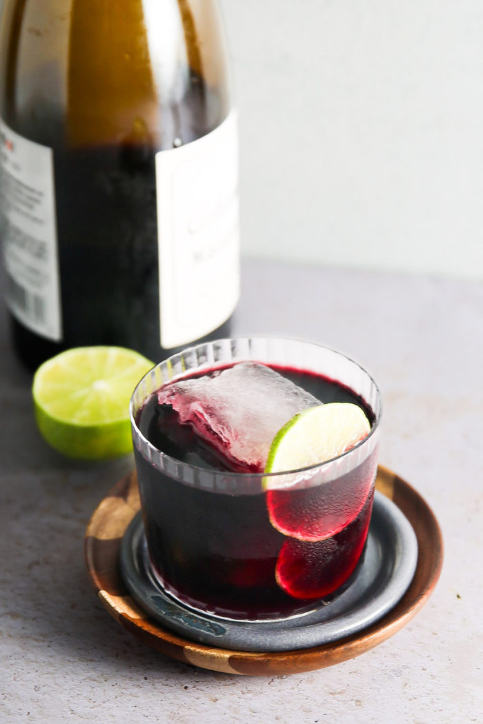 A glass filled with red wine, garnished with a lime slice and a large ice cube, sits atop a wooden and ceramic coaster. Behind the glass is a bottle of Tinto de Verano with an attached label, and a halved lime is placed next to the bottle on a light gray surface.