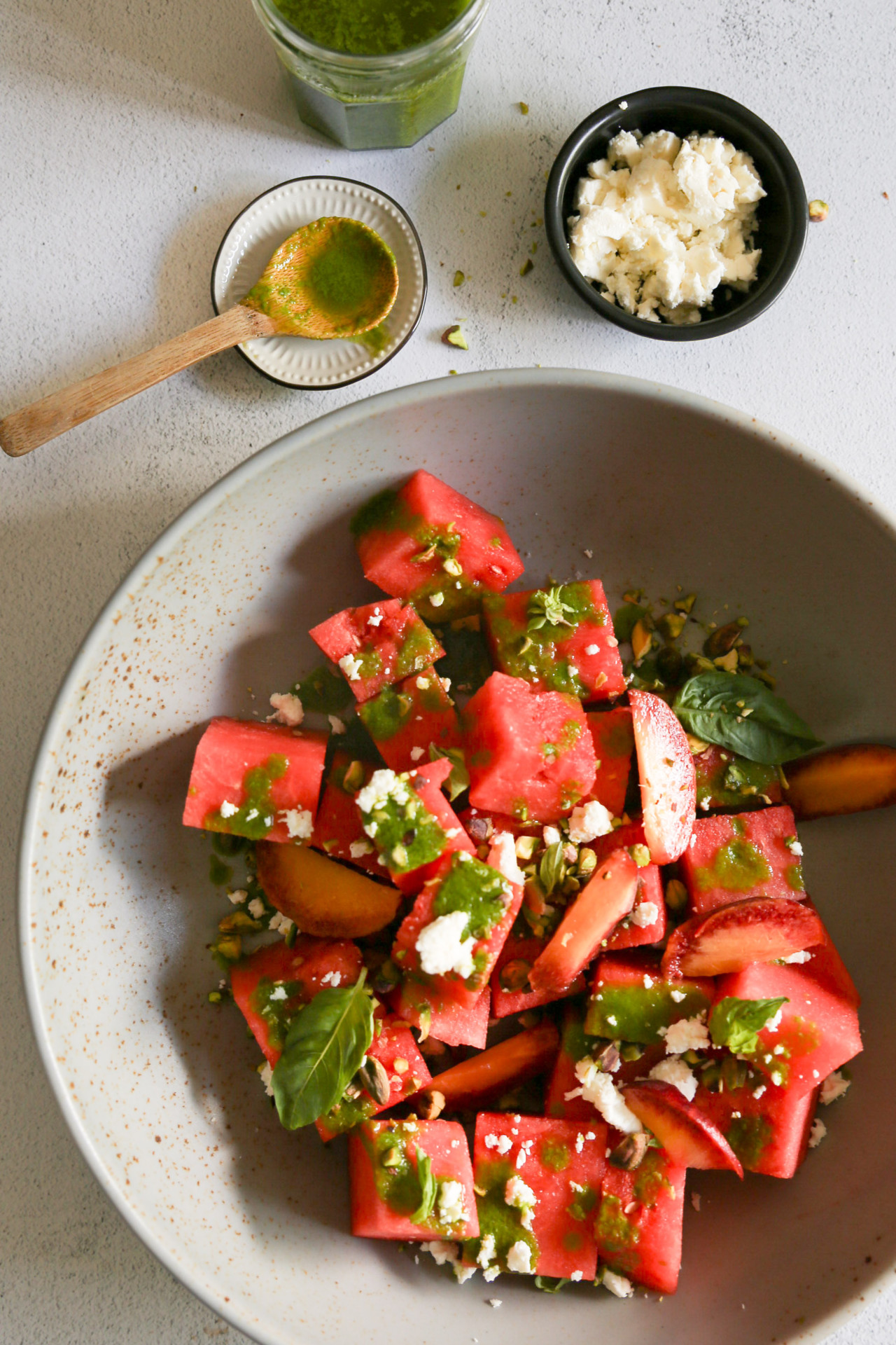 A bowl of watermelon salad with pieces of watermelon and peach, sprinkled with crumbled feta cheese, basil leaves, and a basil vinaigrette. Nearby, a small dish with green powder and a wooden spoon, a black bowl of crumbled feta, and a jar of basil vinaigrette are arranged on the gray surface.