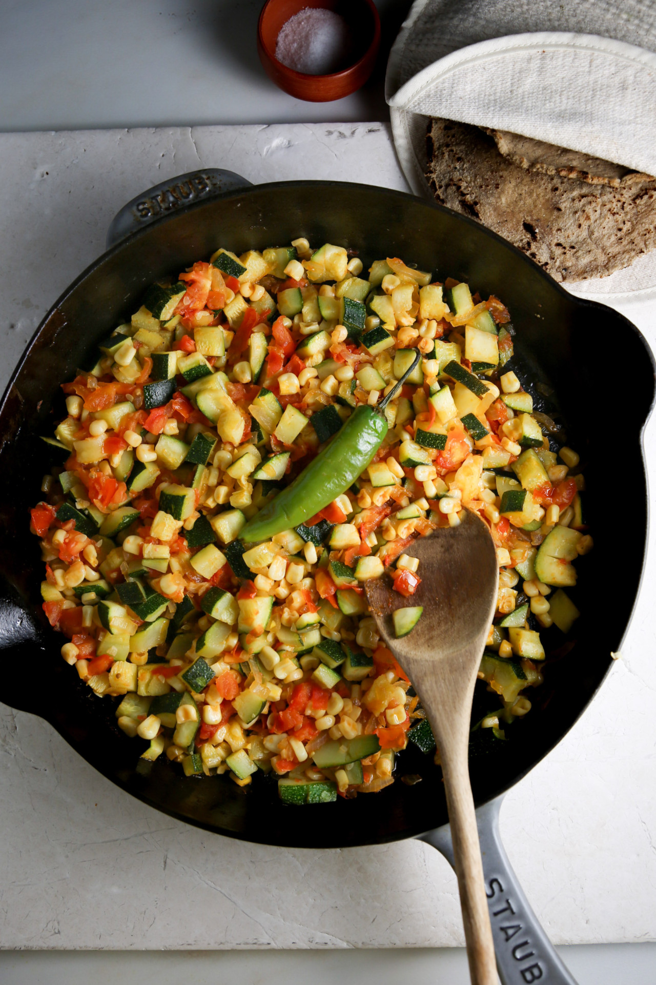 A black Staub pan sits on a white countertop, filled with a colorful mix of chopped vegetables including zucchini, tomatoes, and corn. A green chili pepper rests on top of the vegetables, and a wooden spoon lies in the pan. In the background, there is a small bowl of salt and a stack of tortillas.