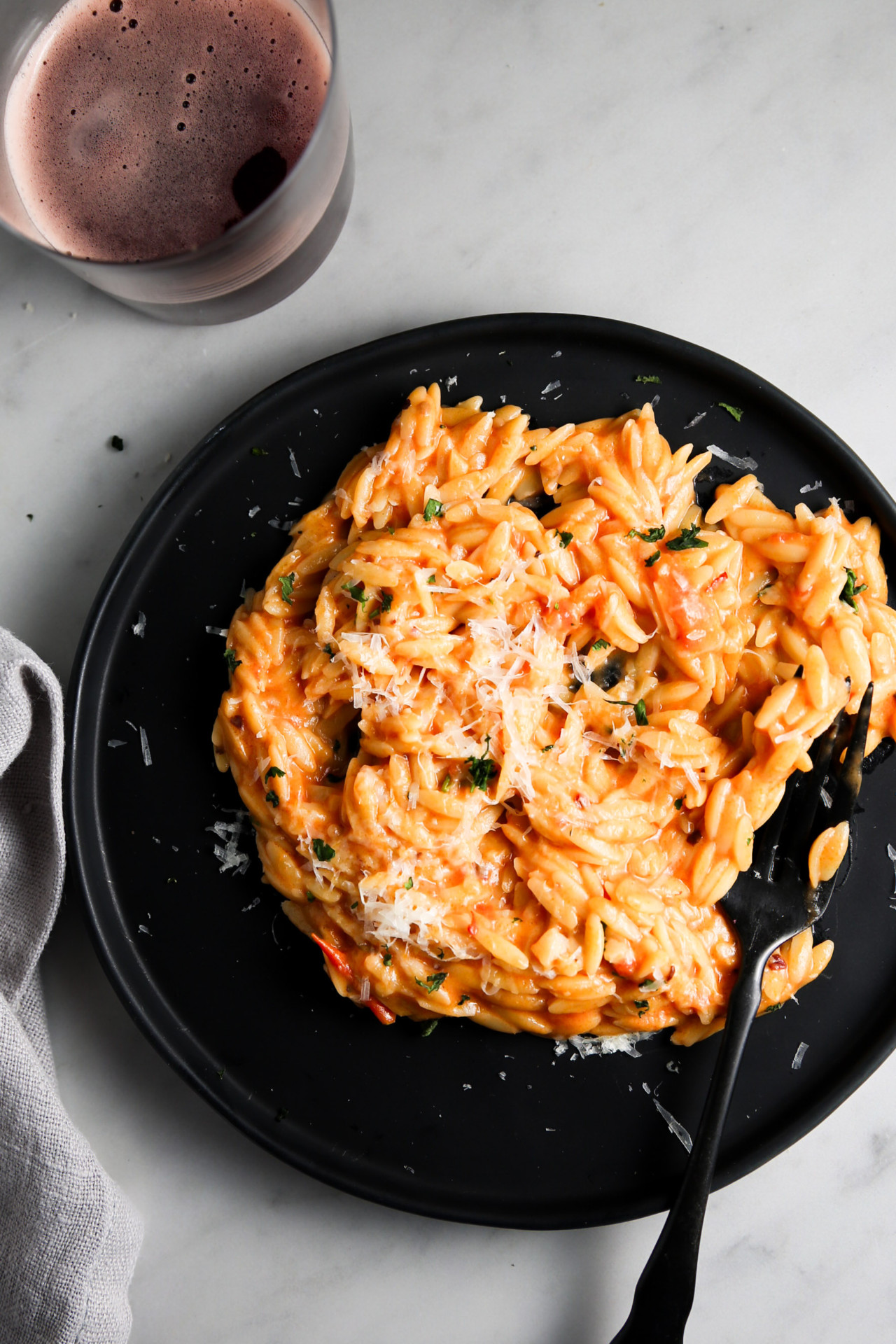 A black plate with creamy orzo pasta topped with grated cheese and herbs. The tomato-based sauce has a touch of spicy flavor, garnished with chopped parsley. A black fork lies on the plate, and a glass of red wine is partially visible at the top left corner of the image. A light gray cloth is placed to the left.