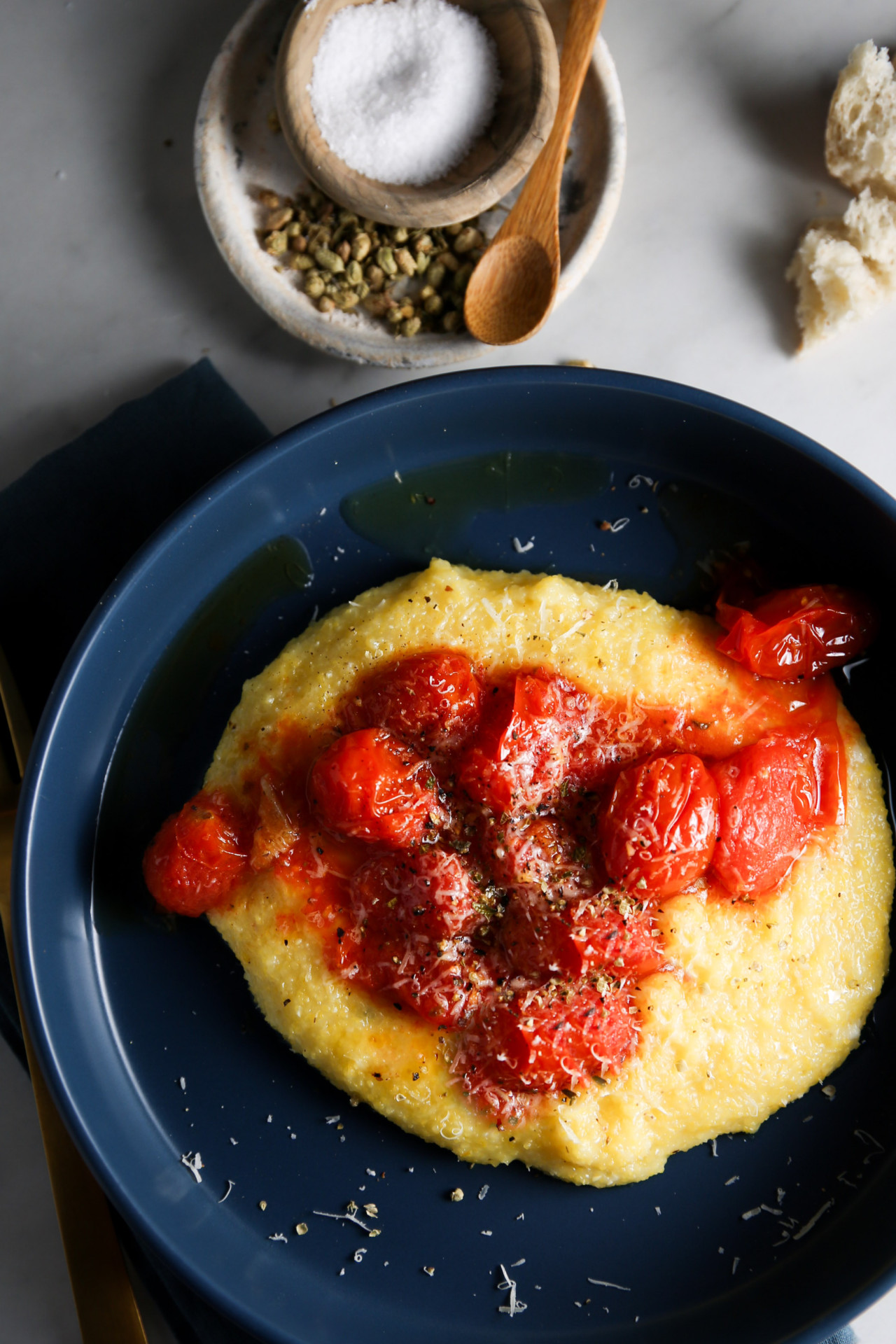 A blue bowl holds a serving of creamy polenta topped with blistered cherry tomatoes and a sprinkle of grated cheese and herbs. Next to the bowl are a small dish of salt with a wooden spoon and a few pieces of crusty bread on a marble surface.