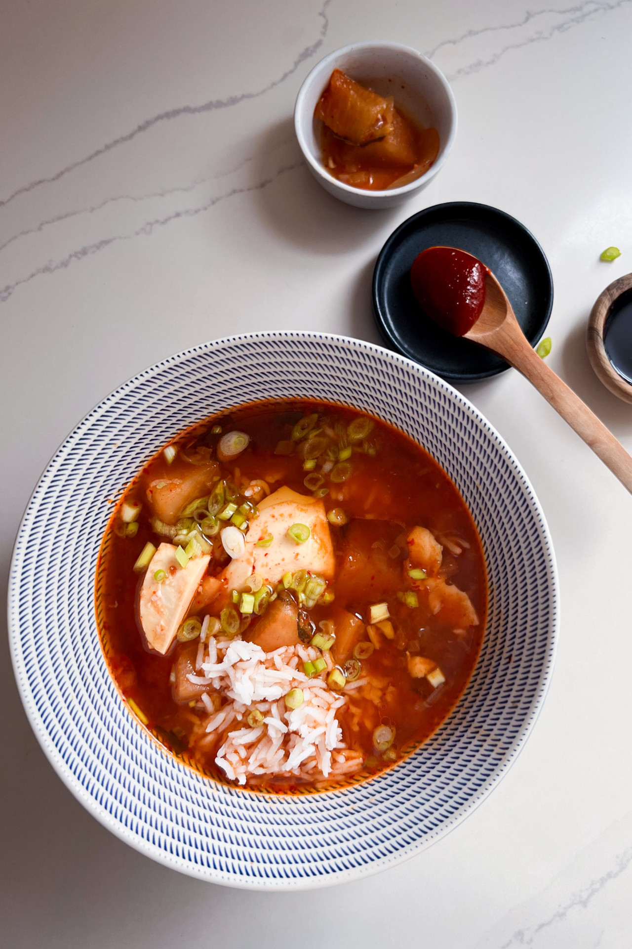 A bowl of vibrant red kimchi stew with tofu cubes, rice, and green onions is placed on a white surface. Beside the bowl are two small dishes: one with a piece of kimchi and the other with a dollop of red paste on a wooden spoon resting on the edge.