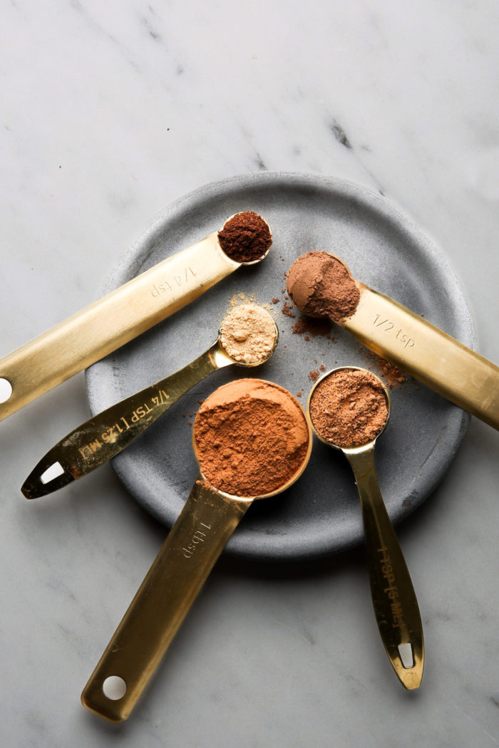 A set of five brass measuring spoons, akin to an artist’s auto draft, is elegantly filled with various spices on a round, gray plate. Labeled precisely from 1/4 tsp to 1 tbsp, the spices present a palette of browns and reds against a marble background.