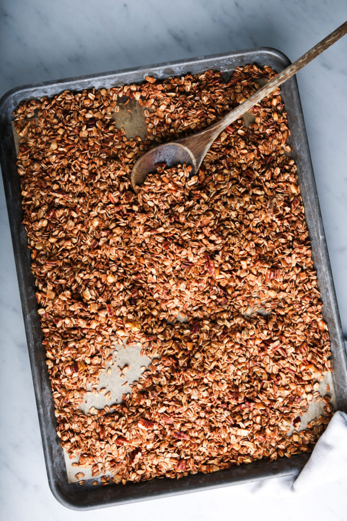 A large baking sheet filled with baked granola sits on a marble counter. The granola is golden brown and textured, with visible oats and nuts. A wooden spoon rests on the granola, suggesting it has been recently stirred. In the corner, a white cloth is slightly visible.