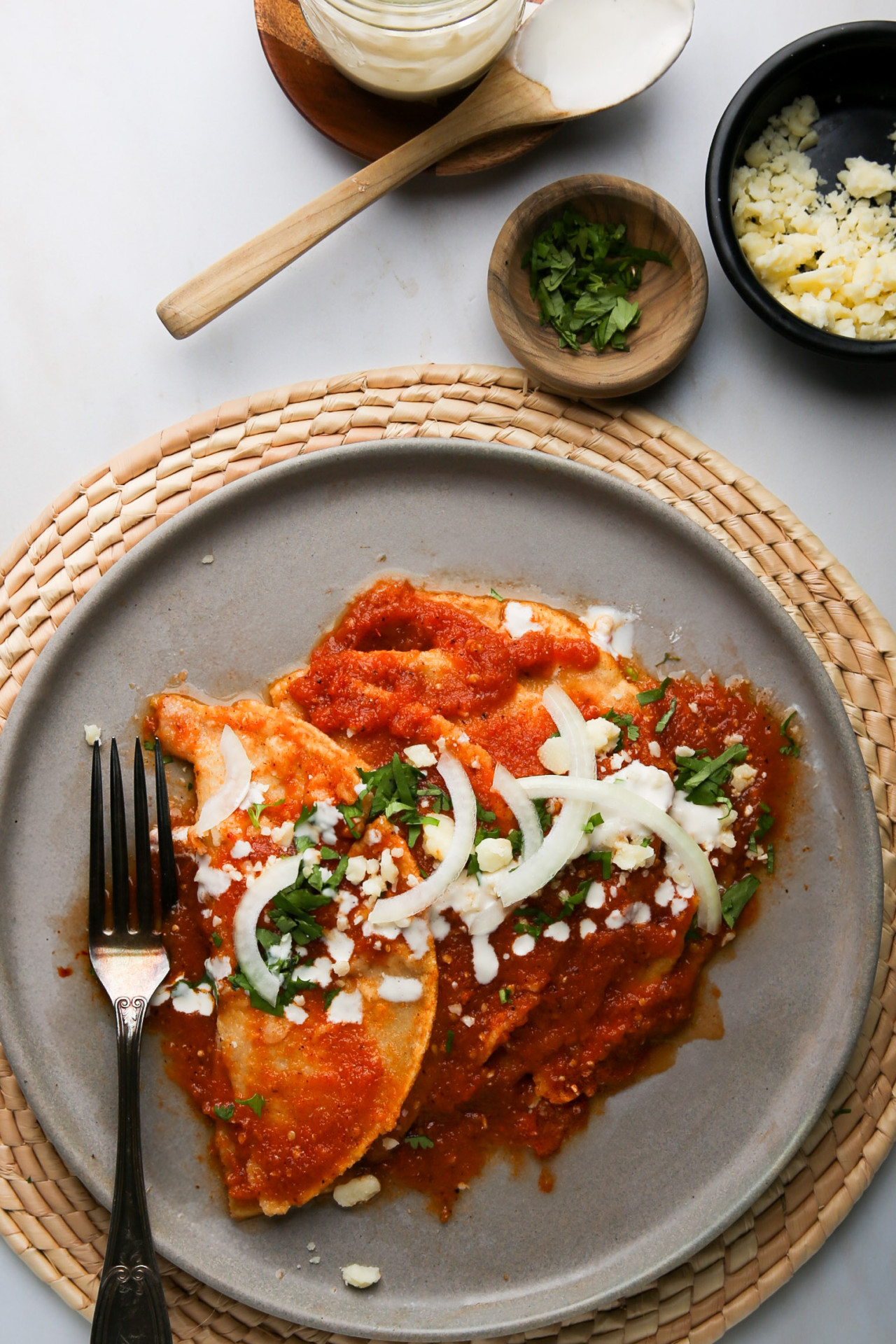 A gray plate on a woven mat showcases Mexican cuisine at its finest: entomatadas drenched in red sauce, garnished with crumbled cheese, sliced onions, and chopped cilantro. A fork lies beside them. Above, a small bowl of sauce with a spoon sits alongside wooden bowls of fresh cilantro and cheese.