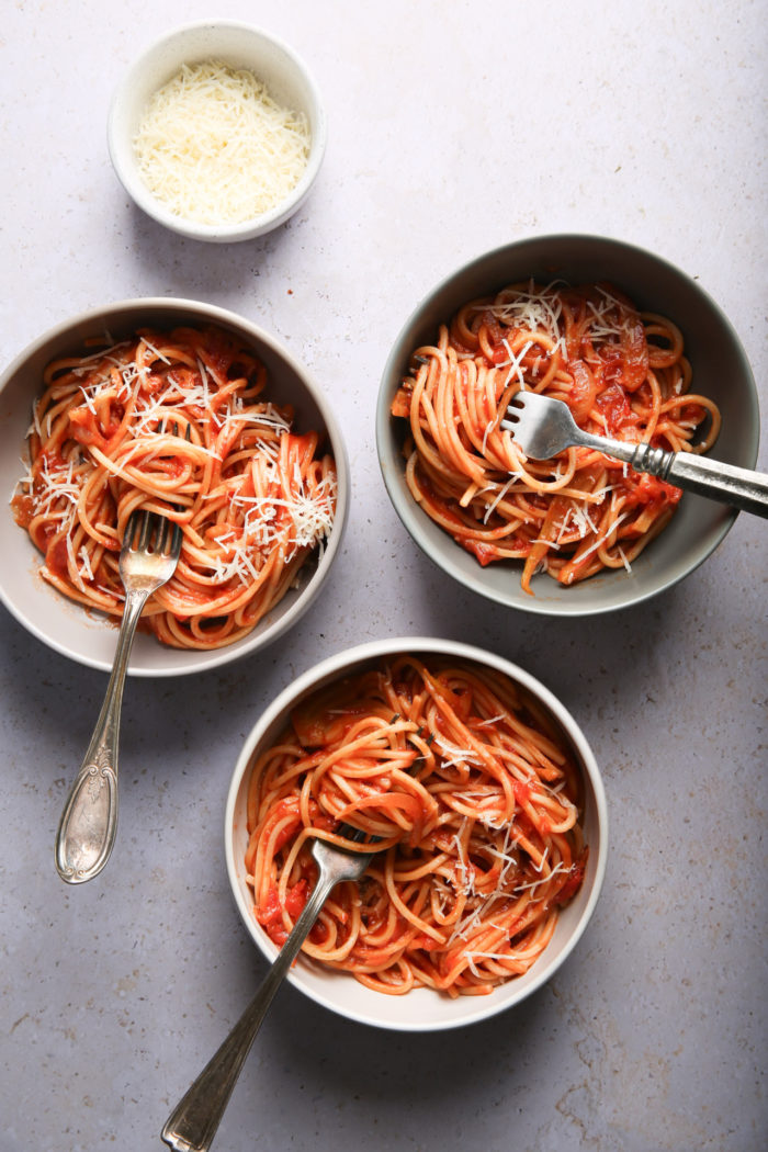 On a light surface, three bowls of spaghetti with tomato sauce and grated cheese are arranged. Each bowl contains a fork. Above the bowls, there is a small plate with extra grated cheese, ready to serve. The well-lit scene highlights the textures of the pasta, and the fennel seeds add an aromatic touch.