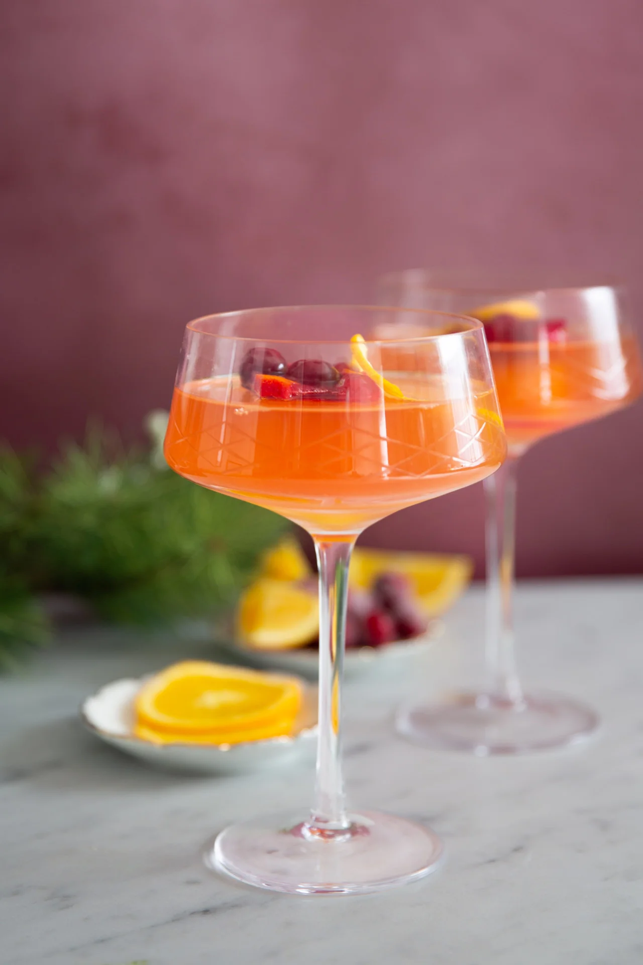 Dos elegantes copas de cóctel llenas de una vibrante y festiva mimosa están decoradas con arándanos y cáscaras de limón. Se encuentran sobre una superficie de mármol con rodajas de naranja y arándanos dispersos en el fondo. El entorno presenta un fondo de vegetación difuminada, que agrega un toque de elegancia natural.